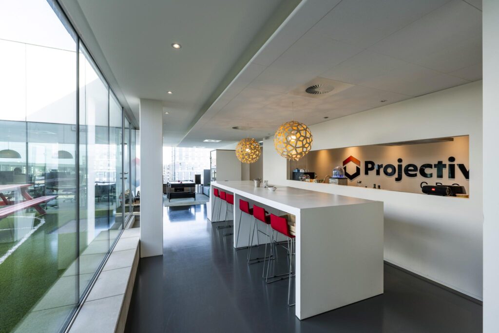 Modern office kitchen area at Projectiv on Krijn Taconiskade with a white island, red bar stools, and decorative pendant lights.
